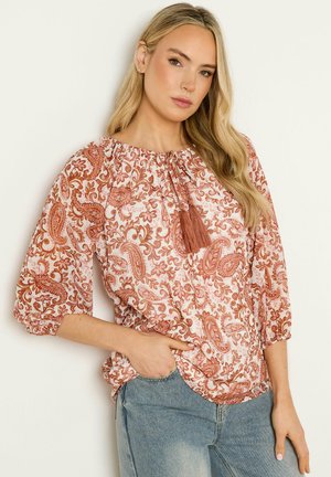 Lyshåret kvinde iført en løs bluse med paisley-mønster og kvaster samt blå jeans, stående med den ene hånd i lommen mod en ensfarvet baggrund.