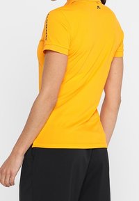 Kortärmad polo i ljusgul tyg med en slät textur, med krage och logotypdetalj på axeln. Svarta shorts syns nedanför.