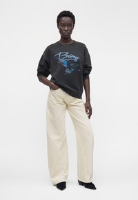 Femme portant un sweat-shirt noir oversize avec un graphisme d'aigle bleu et des lettres, pantalon large crème et chaussures noires pointues, debout avec assurance.