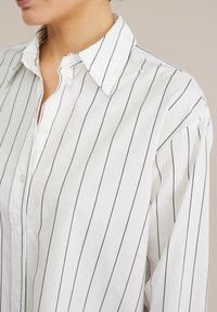 White button-up shirt with black vertical stripes, featuring a pointed collar and loose fit. Smooth fabric with no visible patterns or accents.