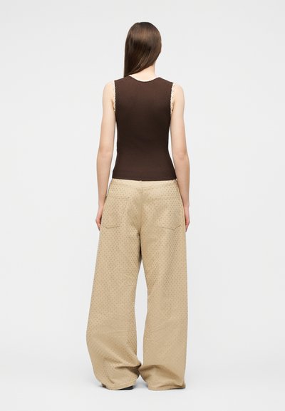 Γυναίκα με μακριά ίσια μαλλιά που φοράει αμάνικη καφέ ριγωτή μπλούζα και φαρδιά μπεζ παντελόνια με διακριτικό πουά μοτίβο, στεκόμενη με την πλάτη προς το μέρος μας.