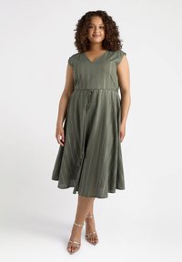 Grünes, knielanges Kleid mit kurzen Ärmeln, V-Ausschnitt und tailliertem Bund. Verfügt über vertikale Streifen und einen strukturierten Stoff. Kombiniert mit silbernen Sandalen.