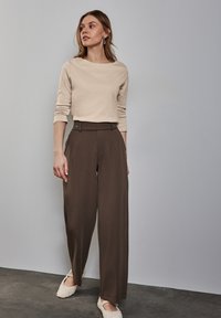 Beige Langarmoberteil mit Rundhalsausschnitt, kombiniert mit hochgeschnittenen braunen weit geschnittenen Hosen mit Knopfdetails. Grauer Hintergrund und dunkler Boden.