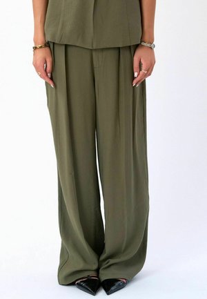 Kobieta ubrana w luźne, oliwkowozielone szerokie spodnie, czarne szpilki z ostrym noskiem, złotą bransoletkę, srebrny zegarek oraz pierścionki na palcach na białym tle.