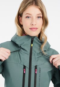 Grüne wasserdichte Jacke mit hohem Kragen, schwarzen Reißverschlüssen und pinken Zuglaschen. Verfügt über eine verstellbare Kapuze und ein kontrastierendes Innenfutter.