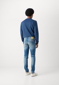 Marineblaues Sweatshirt mit langen Ärmeln, kombiniert mit hellblauen Jeans und einem gelben Etikett auf der Gesäßtasche sowie weißen Sneakern.