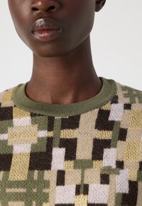 Pull en tricot présentant un motif géométrique en vert, marron et crème, avec un col côtelé vert olive et un petit logo.