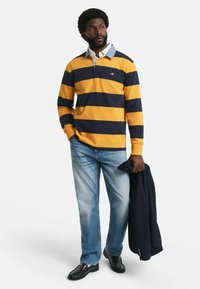 Man in een gele en marineblauwe rugbyshirt, lichtblauwe jeans, zwarte schoenen, houdend een marineblauwe blazer in zijn linkerhand, staand op een witte achtergrond.