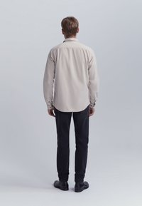 Hombre de pie de espaldas, vistiendo una camisa de manga larga beige, pantalones negros y zapatos negros, frente a un fondo gris liso.