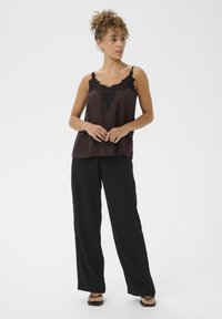 Zwarte camisole met kantafwerking bij de halslijn, gelaagd over zwarte wijde broek. Model draagt zwarte slippers, met een neutrale achtergrond.