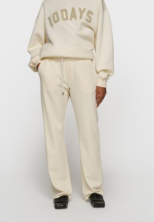 Osoba ubrana w kremowe spodnie dresowe i pasującą bluzę z napisem "10DAYS", do tego czarne buty na kwadratowym nosku.
