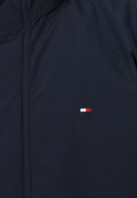 Navy plavi sakou sa malenim izvezenim logom u crvenoj, bijeloj i plavoj boji. Glatka tekstura s skrivenim patentnim zatvaračem i bez vidljivih dijelova.
