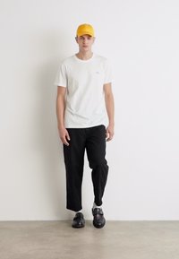 Camiseta blanca de manga corta con un pequeño logo, combinada con pantalones negros ajustados y zapatos negros, complementada por una gorra amarilla brillante.