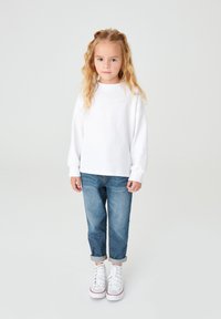 Felpa bianca a maniche lunghe con spalle scese, jeans blu con risvolti e sneaker bianche con dettagli rossi. Il bambino è in piedi su uno sfondo neutro.