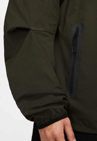 Mörkgrön, lättviktsjacka med detaljer som en zippered ficka på vänster sida och elastiska ärmslut. Slät textur, minimalistisk design.