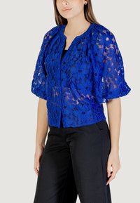 Giacca corta in pizzo blu con motivi floreali, maniche corte a sbuffo e chiusura a bottone. Indossata sopra una maglietta nera e abbinata a pantaloni neri.