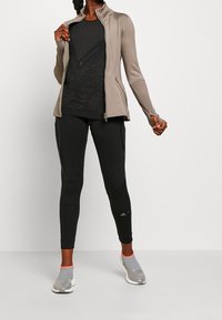 Mujer con chaqueta beige con cremallera, top de manga larga negro con estampado, leggings negros ajustados y zapatillas gris claro sin cordones con ribete rosa.