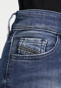 Denim pants with a high waistband, featuring a back pocket with a smaller pocket inside, in a medium blue wash with faded texture.