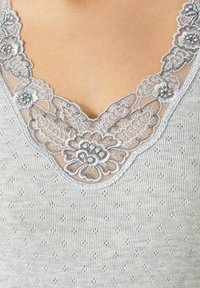 Canotta grigia con pizzo, scollatura a V, caratterizzata da motivi floreali e un tessuto a maglia strutturato con piccoli fori decorativi.