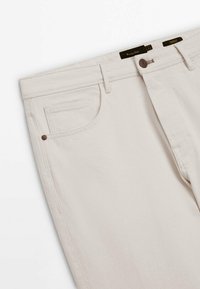 Beiget farkut pehmeästä puuvillasta, joissa on klassinen viiden taskun muotoilu, hienovarainen tekstuuri ja metallinen nappisuljenta ruskeilla yksityiskohdilla.