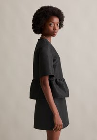 Melnā īsām piedurknēm pepluma topa ar teksturētu audumu; vienkrāsaina saderīga svārku daļa. Modelis pozē sānis pret neitrālu fonu.