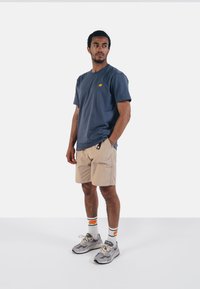 Tmavě modré bavlněné tričko s žlutým logem slona, spárované s béžovými kraťasy. Nosit s šedými teniskami a bílými ponožkami s oranžovými akcenty.