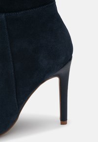 Botte à talon haut en daim bleu marine. Présente une texture lisse, un talon aigu en forme d'éventail effilé, et une couture discrète le long du côté.