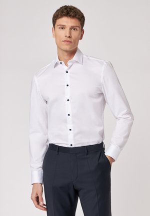 Weißes Hemd mit slim fit, featuring einen Kentkragen, dunklen Knöpfen und langen Ärmeln, kombiniert mit dunklen maßgeschneiderten Hosen.