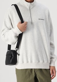 Sudadera gris con cierre de cuarto y marcas negras, combinada con un pequeño bolso cruzado negro que cuenta con cremallera y correa ajustable.