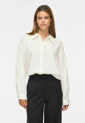 Femme aux cheveux bruns et lisses porte une chemise blanche ample boutonnée, rentrée dans un pantalon noir taille haute, debout sur un fond uni.