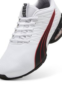 Weißer Sportschuh mit Obermaterial aus Mesh, schwarzen und roten Akzenten sowie strukturierten Details. Verfügt über ein Schnürsystem und eine gepolsterte Sohle.