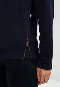 Top de manga larga azul marino con cremallera lateral de latón parcialmente desabrochada cerca de la cadera, usado con pantalones oscuros, mano descansando al lado del muslo.