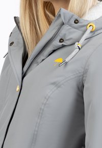 Grå vattentät jacka med dragkedja framtill, huva och vit dragsko. Har en gul paraply-logotyp och silverdetaljer.