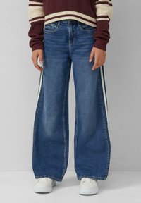 Jeans a gamba larga in denim blu medio con strisce laterali bianche. Il tessuto ha una leggera lucentezza, con un design tradizionale a cinque tasche.