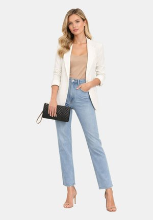 Donna che indossa un blazer bianco, top beige, jeans azzurri chiari, tacchi alti beige, tenendo una pochette nera trapuntata.