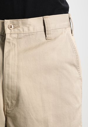 Pantalones de algodón beige con corte recto, que cuentan con cierre de botón y trabillas para cinturón. La tela tiene una textura suave y un ligero brillo.