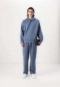 Sweat à capuche bleu décontracté avec une poche avant, pantalon bleu assorti et baskets blanches. Un petit sac noir est porté à la hanche. Texture de tissu douce.
