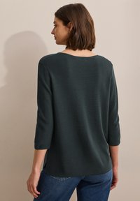 Dunkelgrünes, strukturiertes Strickoberteil mit Dreiviertelärmeln und weitem Ausschnitt, von hinten vor einer neutralen Wand betrachtet.