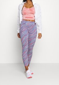 Rosa kamouflage crop top med "PUMA"-logotyp, vit lättvikts zip-up jacka och lila leggings med vågiga rosa och vita ränder, tillsammans med vita sneakers.