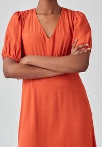 Robe orange avec un décolleté en V profond, manches courtes bouffantes et texture lisse. Le tissu est léger et tombe doucement.
