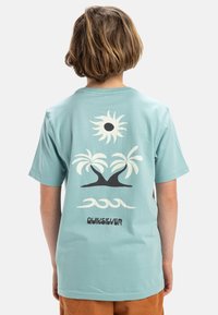 Camiseta de algodón azul claro con un gráfico de un sol, palmeras, olas y el nombre de la marca "Quiksilver" en negro. Mangas cortas y cuello redondo.