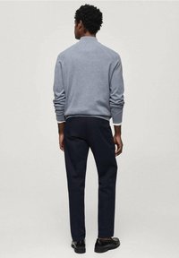 Grauer Pullover mit hohem Kragen, dunkelblaue Hose und schwarze Schuhe; glatte Textur und schlanke Passform mit dezenten Nahtdetails.