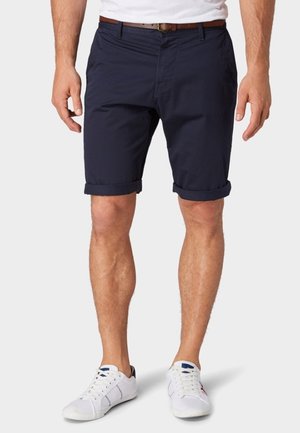 Tummansiniset puuvillashortsit, joissa on käänteet, etutaskut ja vyölenkit, yhdistettyinä valkoisiin tennareihin, joissa on sinisiä ja punaisia yksityiskohtia.