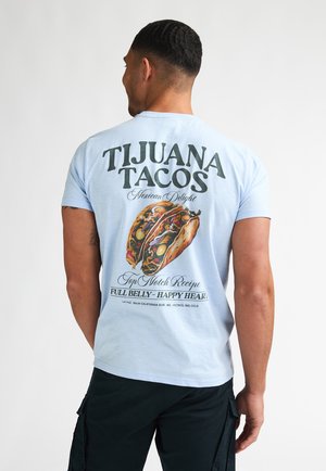 Lichtblauw T-shirt met een grafische print van een taco en tekst: "TIJUANA TACOS Mexican Delight" in kleurrijke, dikke letters.