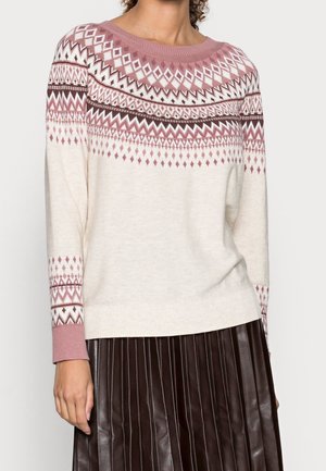 Femme portant un pull en maille crème avec des motifs géométriques rose et marron et une jupe plissée marron foncé.