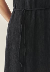 Crna, teksturirana haljina s naboranim uzorkom, koja ima vezanu struk za bolje prianjanje. Tkanina izgleda lagano i fleksibilno.