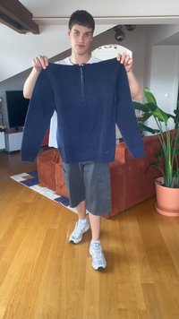 Ung mann som står innendørs og holder en marineblå jakke med glidelås foran seg, iført grå shorts og hvite sneakers.
