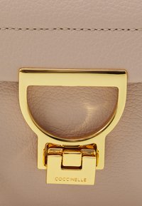 Rosa lærveske med gullfarget lukking og sirkulært håndtak med gravert logo-detalj, som vektlegger et strømlinjeformet design og tekstur.