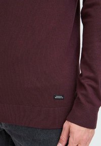 Burgunderrød genser med ribbet tekstur, tettsittende mansjetter og et vevd merke i kanten. Stoffet er mykt og lett.