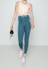 Débardeur de sport rose clair noué à la taille, assorti à un legging taille haute bleu sarcelle foncé avec des accents gris et une ceinture noire.